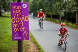 COVID-19 қаупі туралы ескерткен жазудың жанынан өтіп бара жатқан велосипед мінген адамдар. Атланта, АҚШ, 1 шілде 2020 жыл.